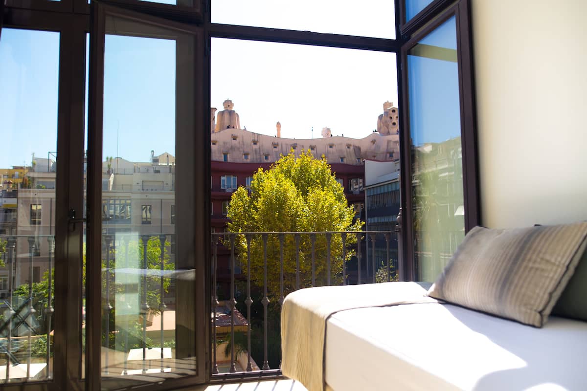 A balcony with a bed, a view of a tree, and buildings in the background.