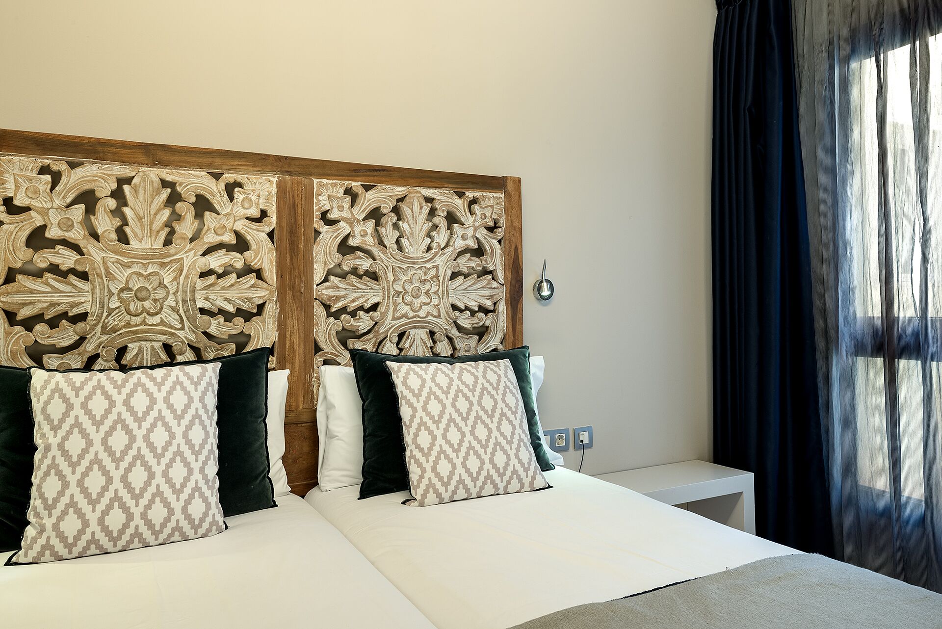 Habitación de hotel con una cama grande, cabecera de madera con detalles tallados, dos mesitas de noche y una ventana con cortinas oscuras.