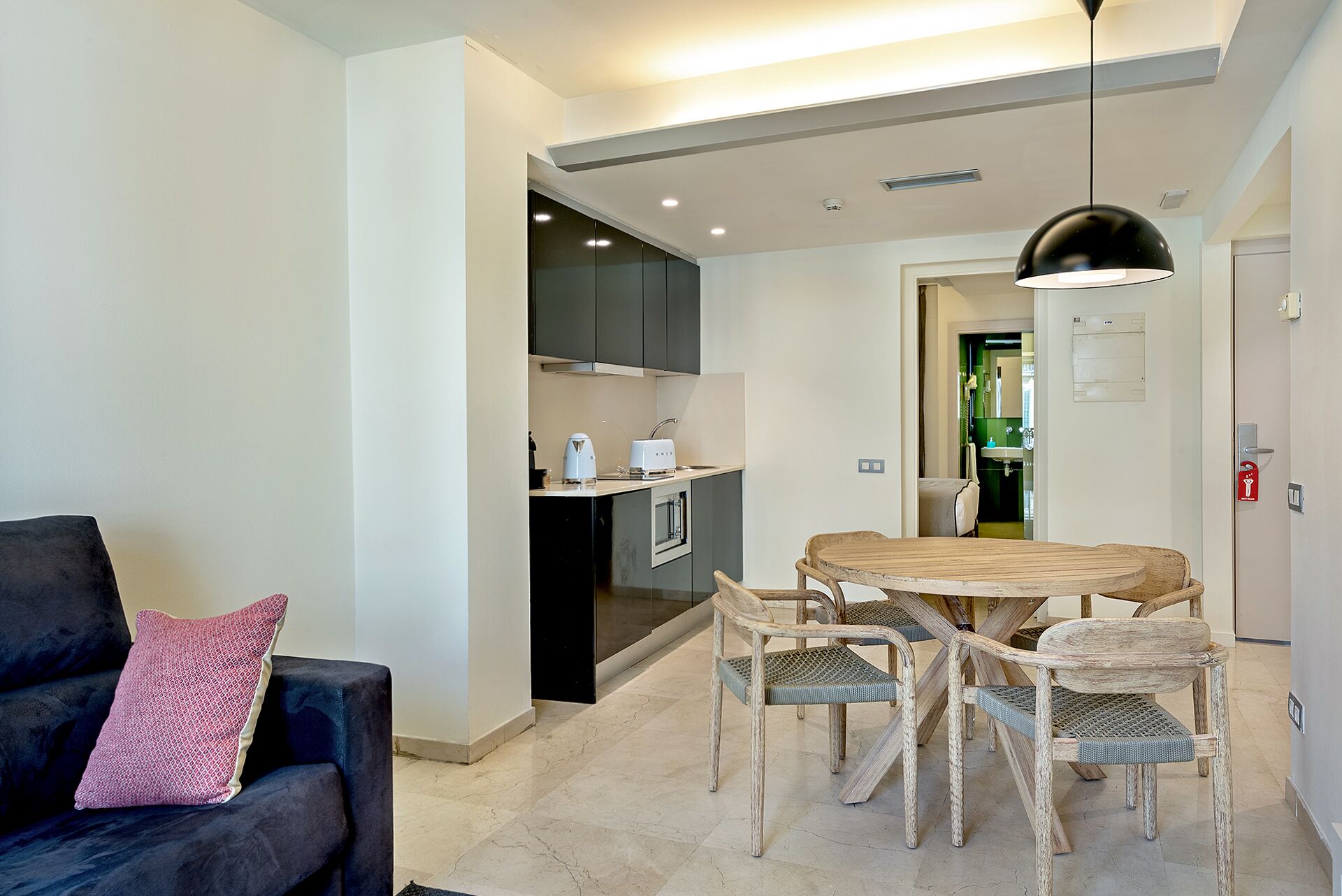 A modern living room with a dark sofa, a wooden dining table with wicker chairs, and a kitchen area with dark cabinets and a white appliance.
