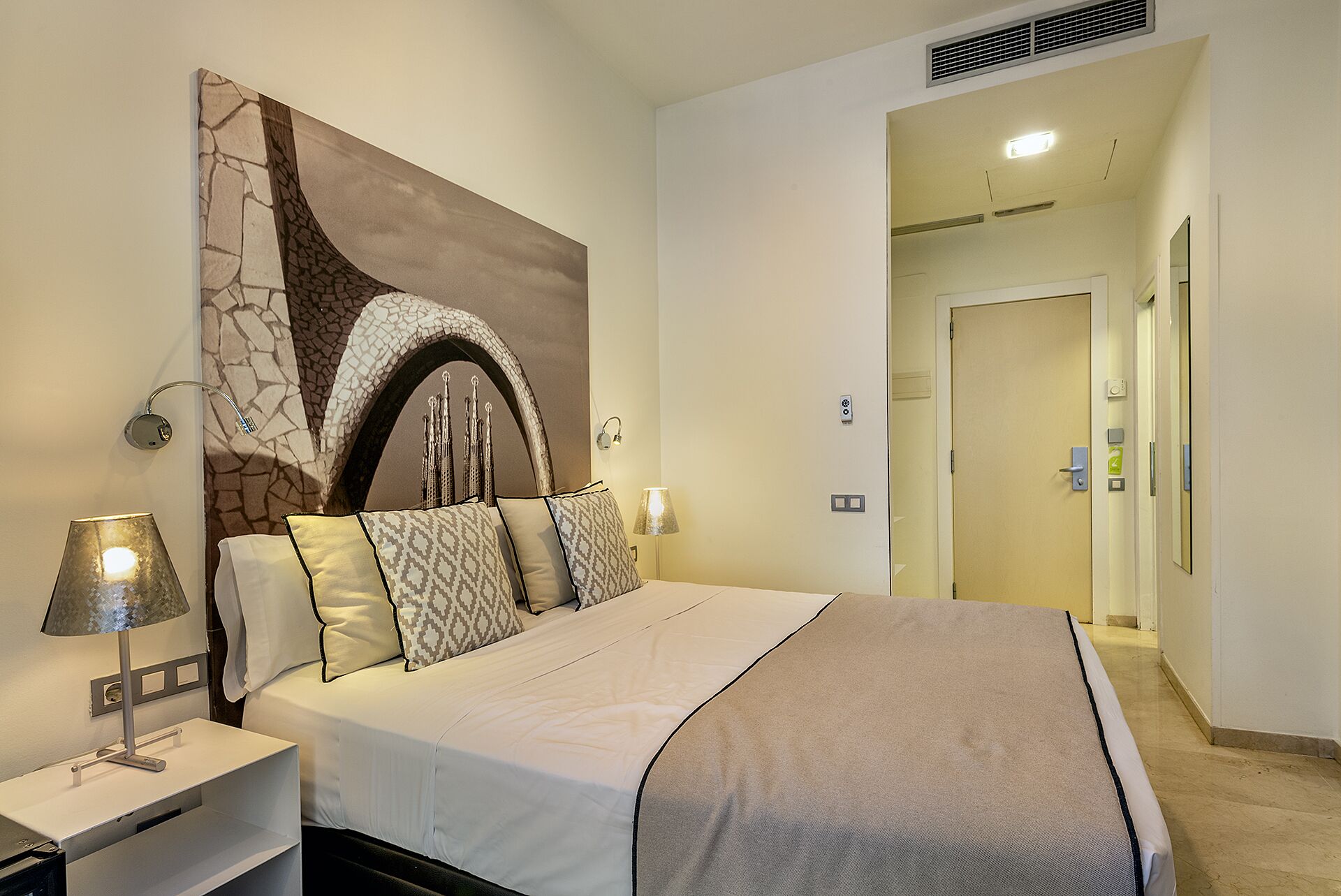 Ein Hotelzimmer mit einem großen Bett, einem Nachttisch mit Lampe, einem Wandbild und einer Tür zu einem weiteren Raum.