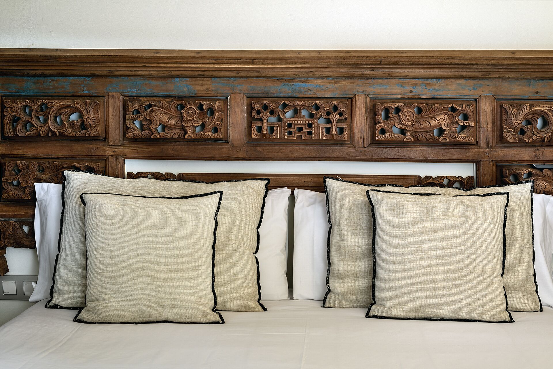 Een bed met een houten hoofdbord, versierd met gedetailleerde houtsnijwerken en vier beige kussens met zwarte bies.