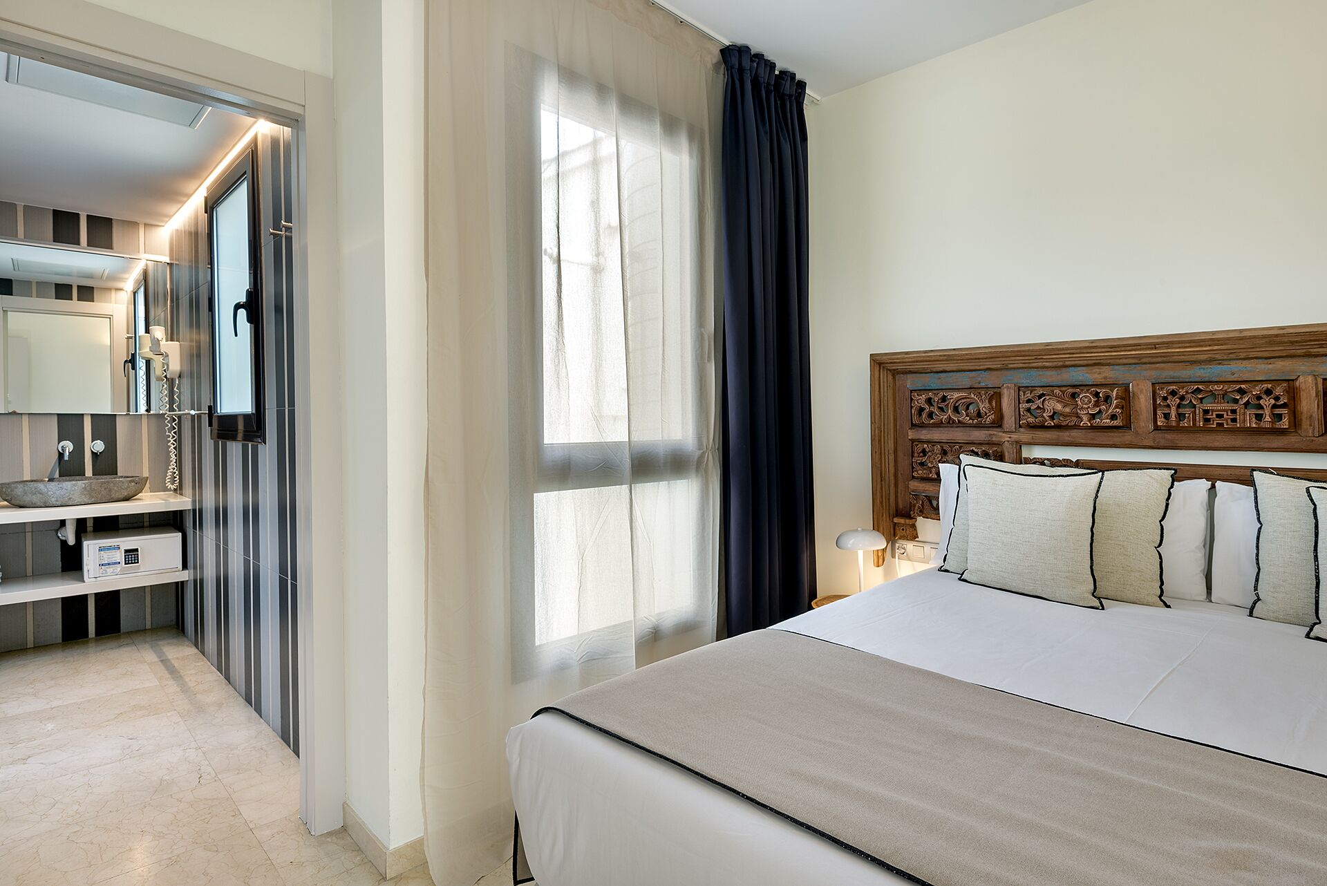 A hotel room with a large bed, a wooden headboard, a bedside table with a lamp, a mirror, a bathroom with a sink and a toilet, and a window with curtains.
