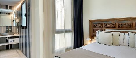 A hotel room with a large bed, a wooden headboard, a bedside table with a lamp, a mirror, a bathroom with a sink and a toilet, and a window with curtains.