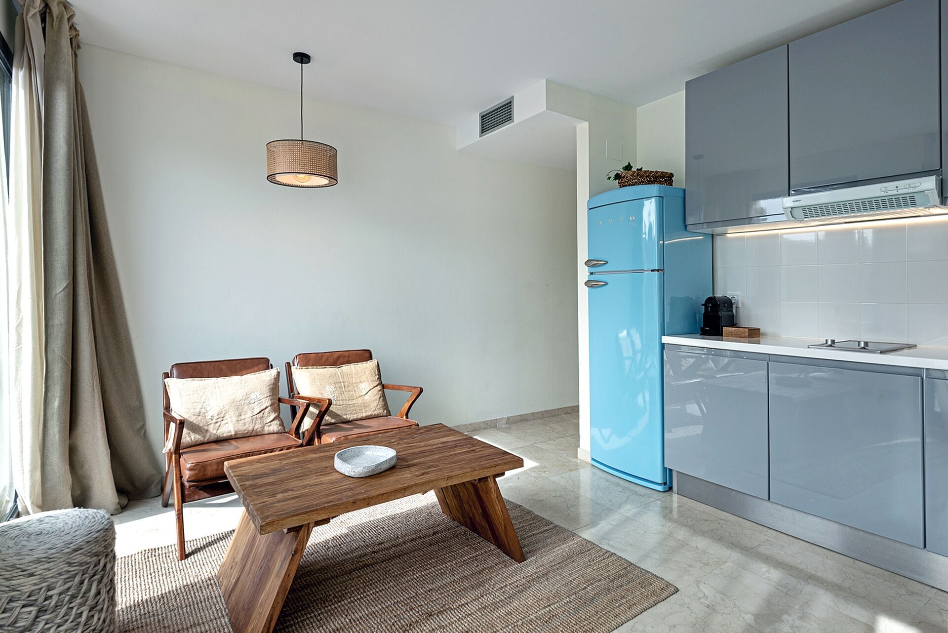 Un espace de vie compact comprenant un coin cuisine, un espace repas avec une table en bois et deux chaises, ainsi qu’un luminaire suspendu.