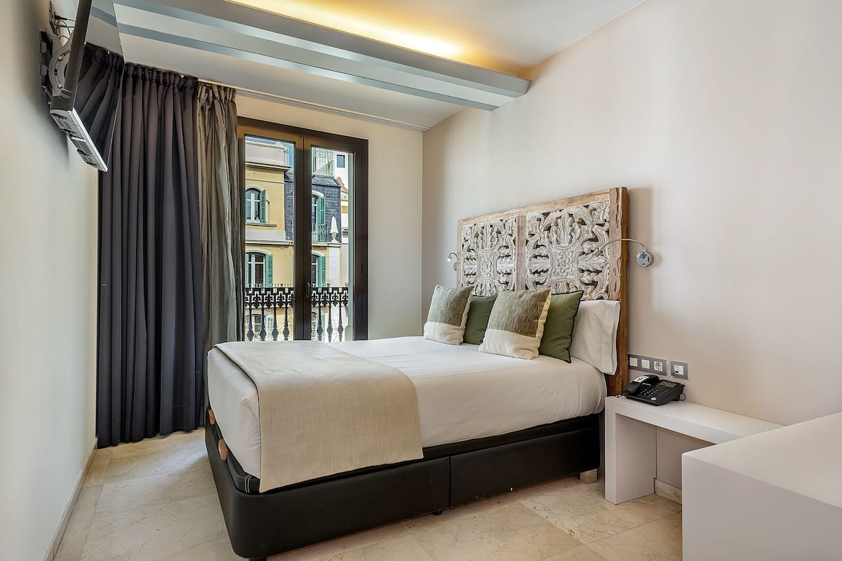 A bedroom with a bed, a desk, and a view of buildings through a window.