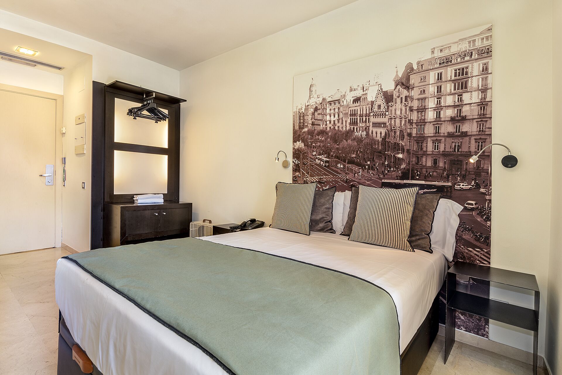 A hotel room with a bed, a nightstand, a dresser, a mirror, and a wall with a cityscape mural.