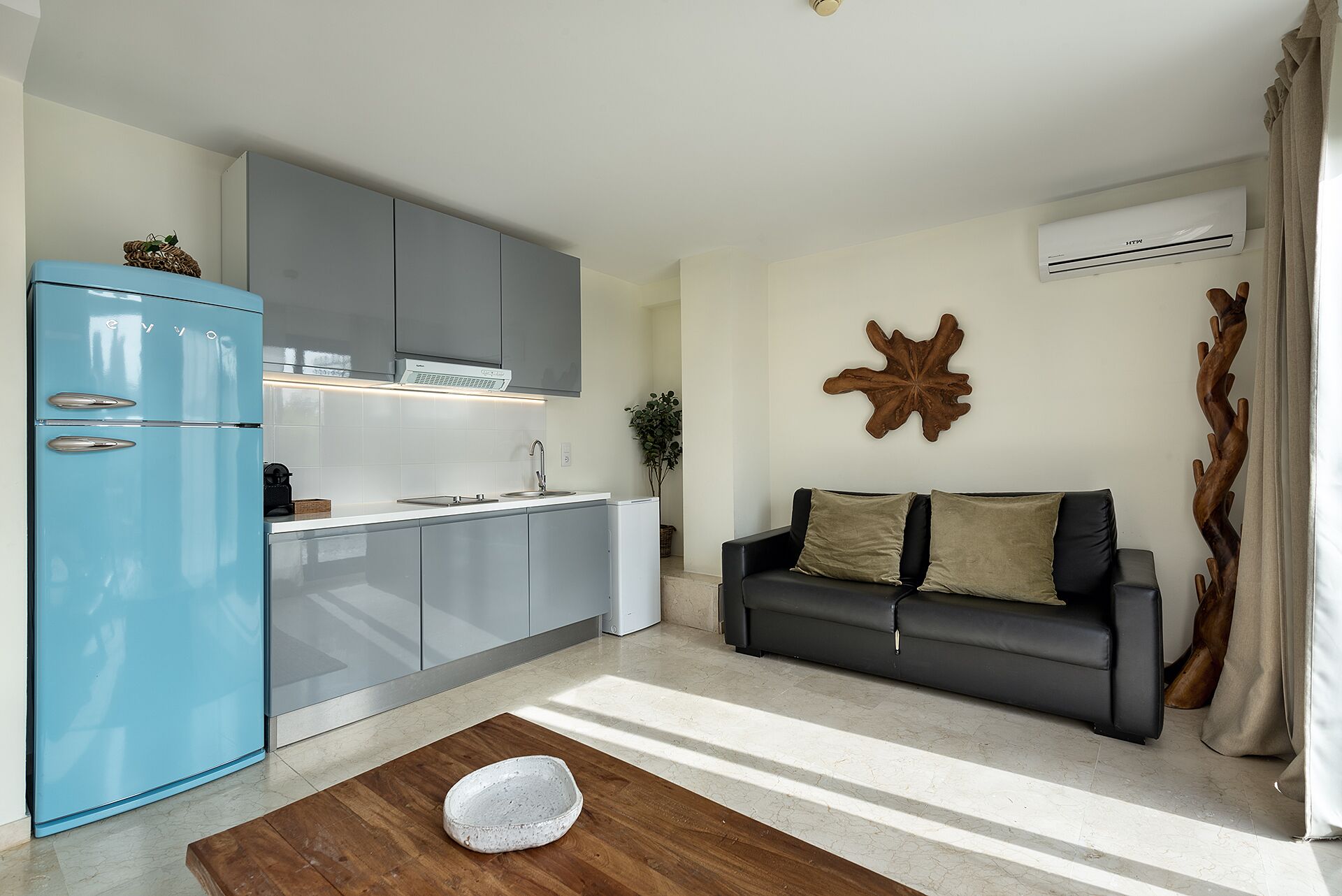 A modern kitchen with grey cabinets and a blue refrigerator, a black sofa with cushions, and a wooden floor.