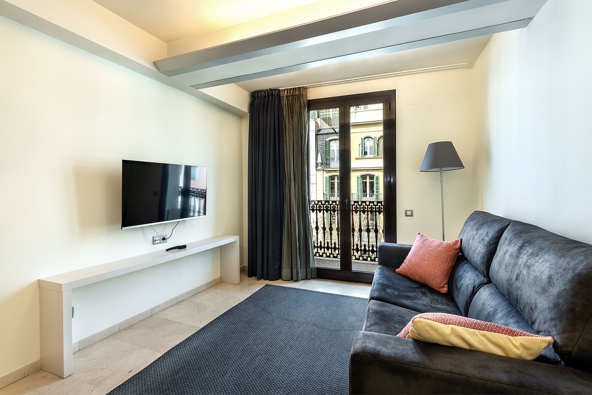 Un soggiorno moderno con una TV a schermo piatto su uno scaffale bianco, un divano grigio scuro con cuscino rosso e giallo, una lampada da terra nera e vista su un edificio tramite una porta di vetro con tenda nera.