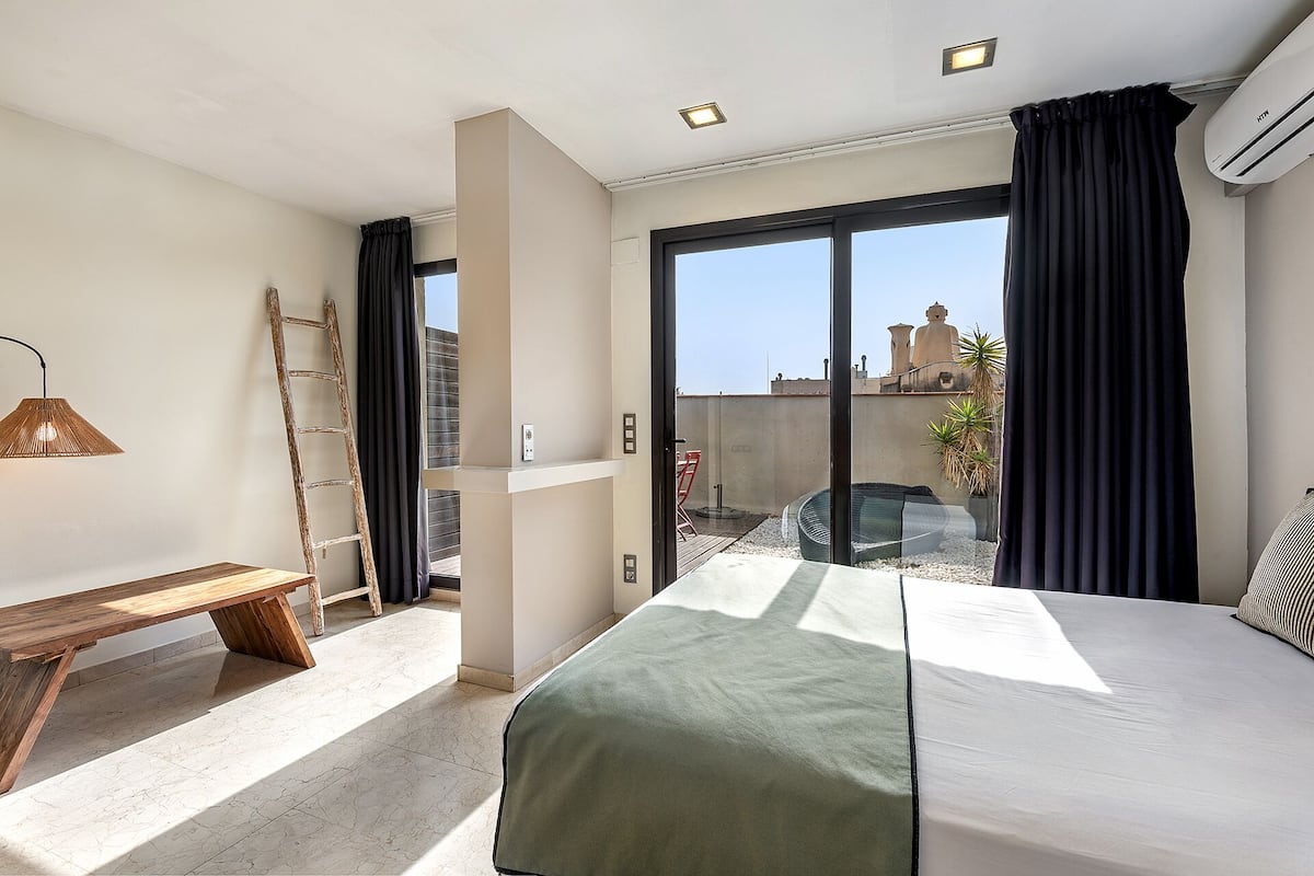 A hotel room with a bed, a bench, a ladder, and a view of the cityscape through large windows.