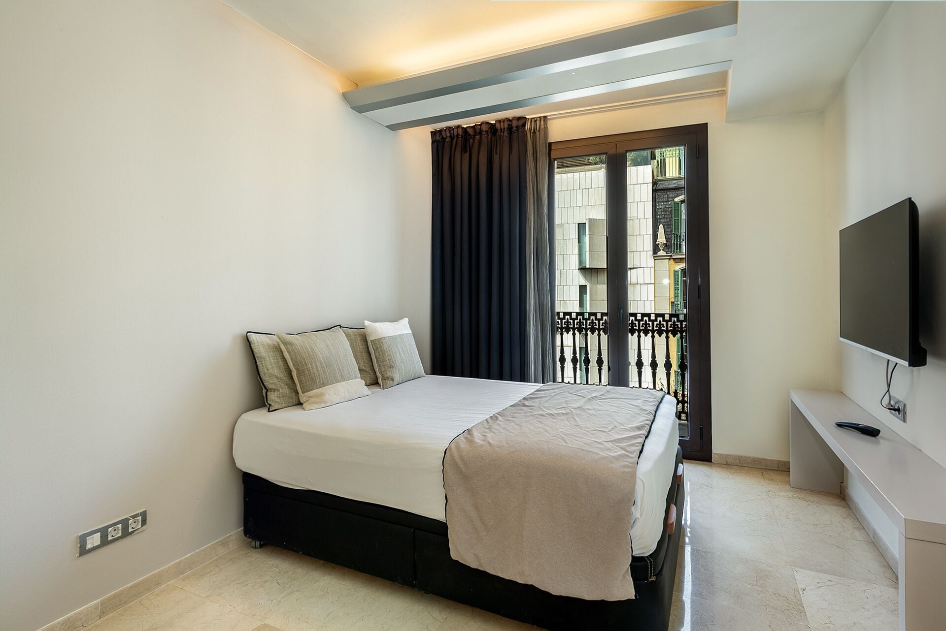 Un dormitorio con cama, televisión y balcón con vistas a edificios.