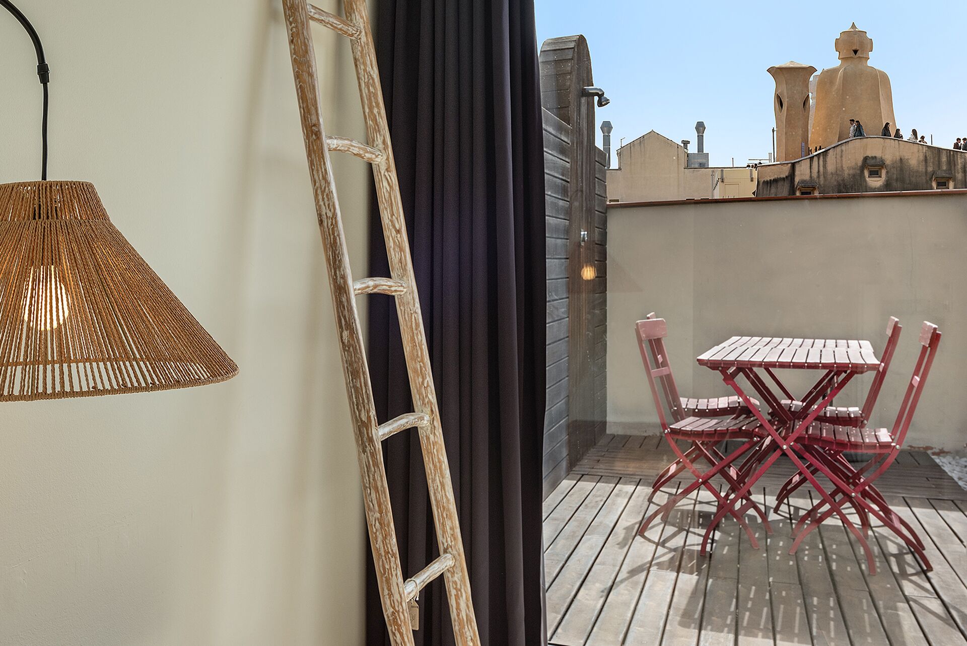 Un balcón con una mesa de madera y sillas rojas, una lámpara de mimbre y vistas a edificios al fondo.