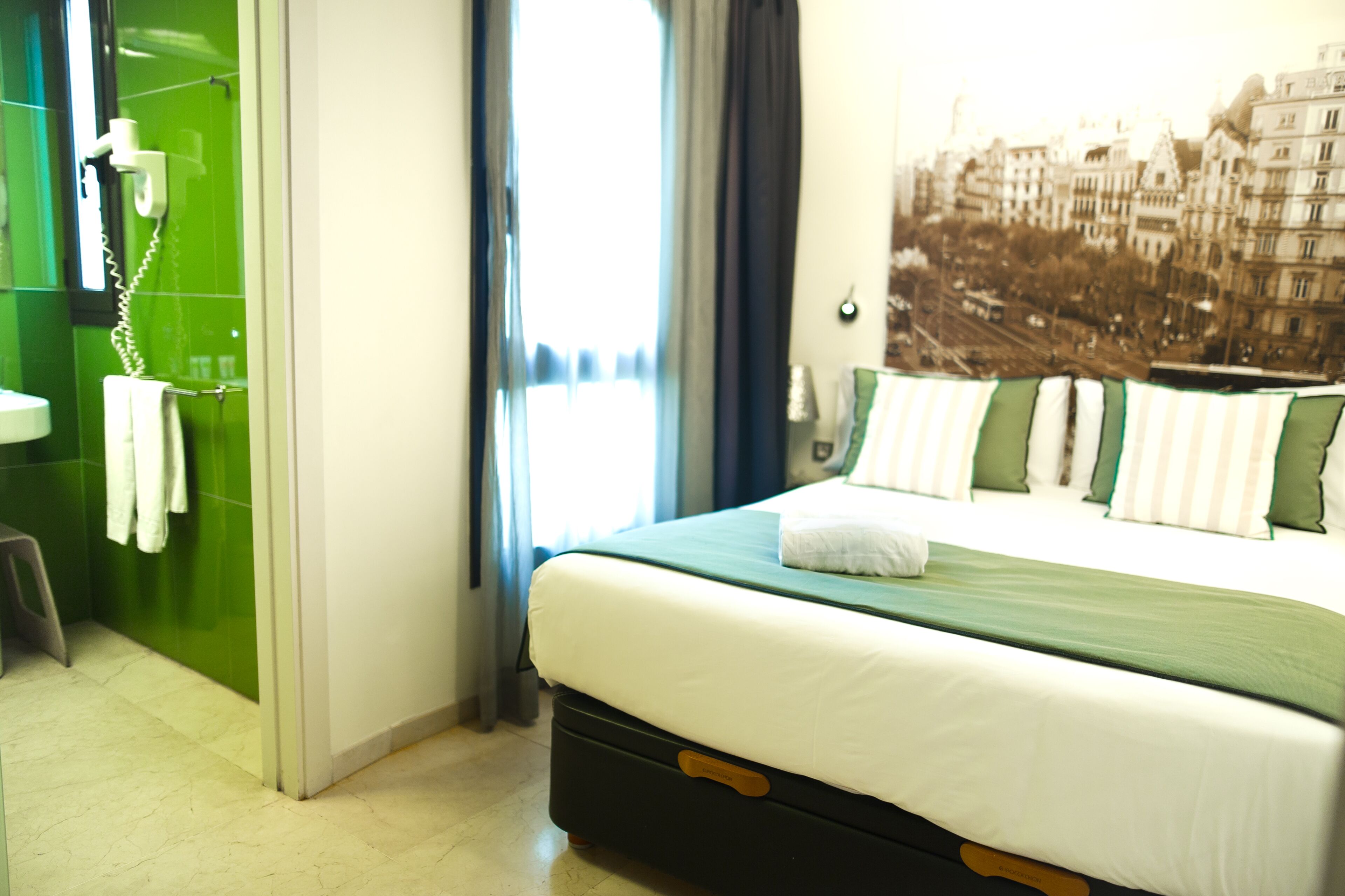 A hotel room with a bed, a bathroom with a green wall, and a cityscape view.