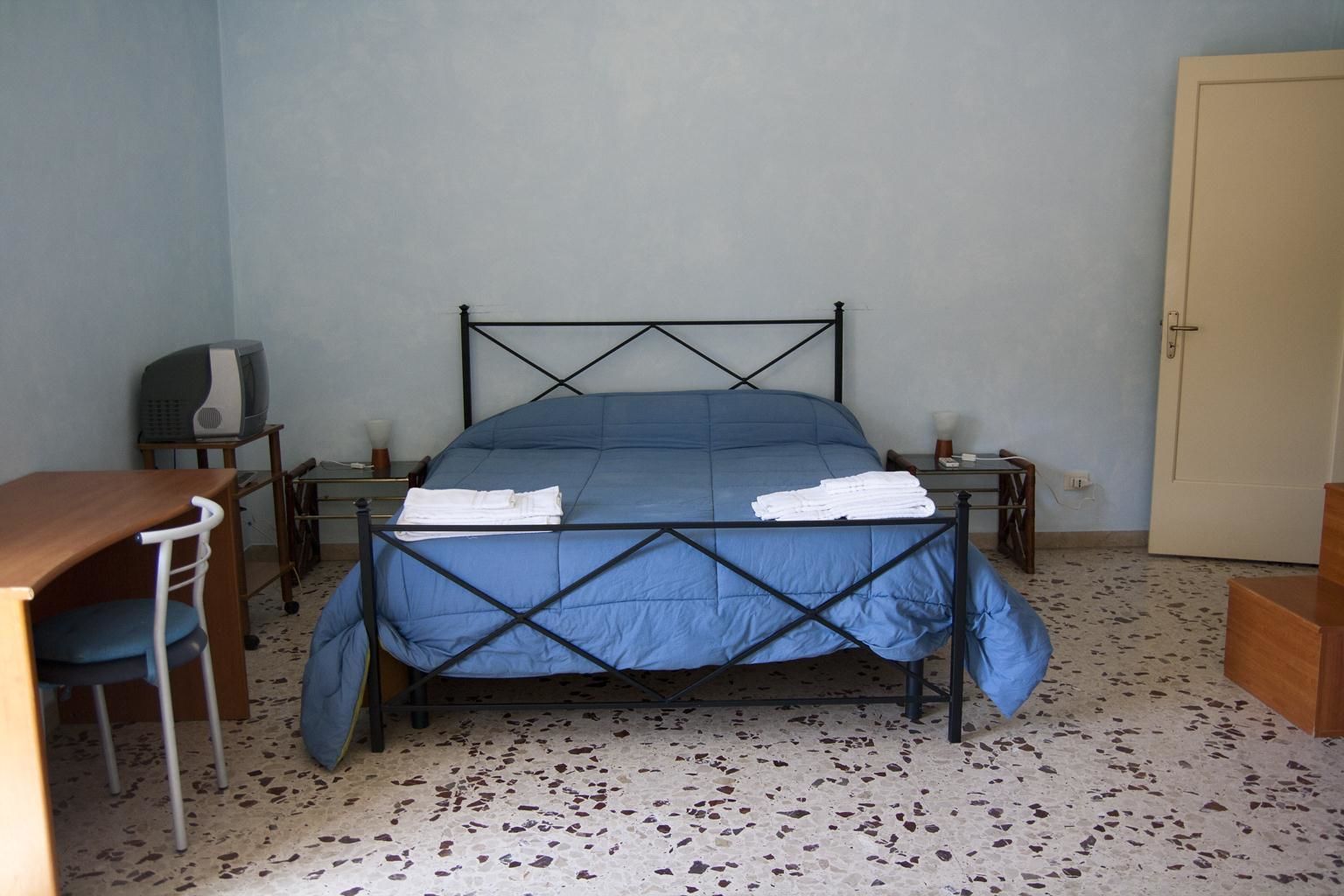 A single bed with a blue bedspread and white towels, a desk with a chair, a television, and a door.