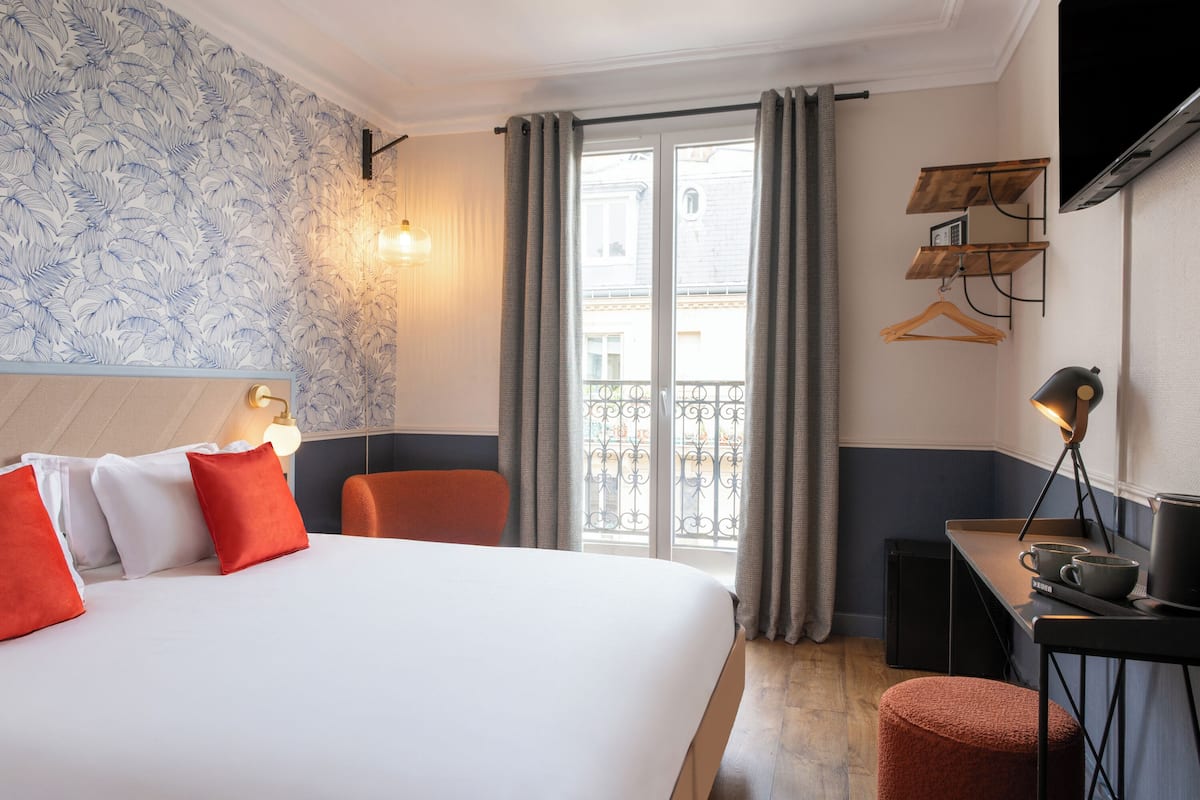 Une chambre d’hôtel avec un lit, un bureau, une chaise, une télévision et un balcon avec des rideaux.