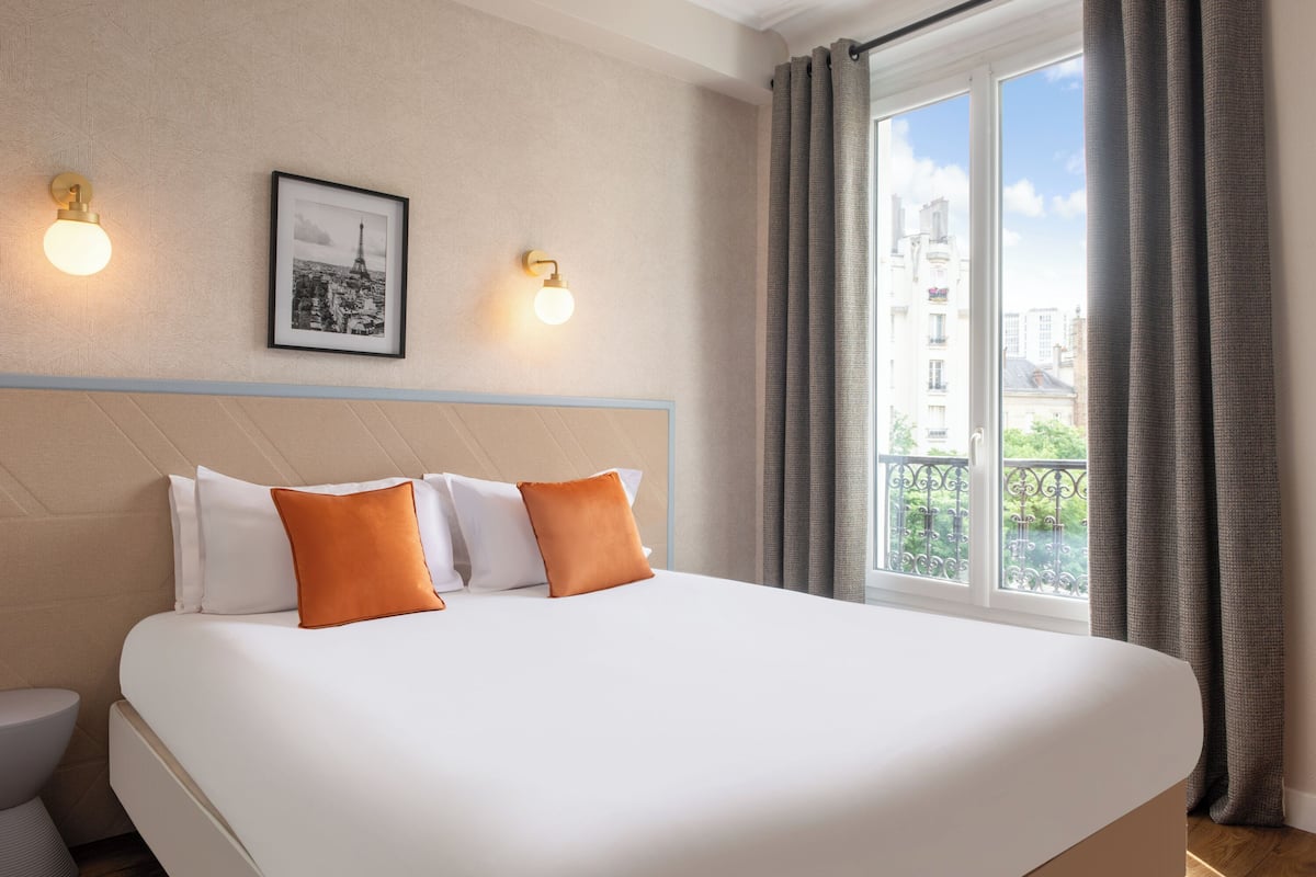 Une chambre d’hôtel avec un grand lit, deux lampes fixées au mur, un tableau encadré et une fenêtre avec des rideaux.