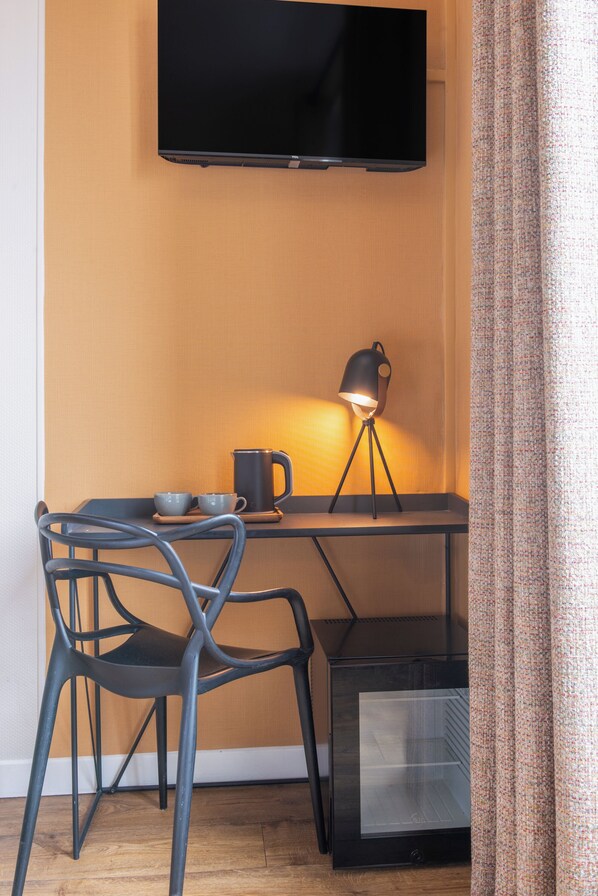 A compact room with a flat-screen TV mounted on the wall, a small table with a lamp, a chair, and a mini-fridge. - Hôtel Coypel by Magna Arbor (Paris)
