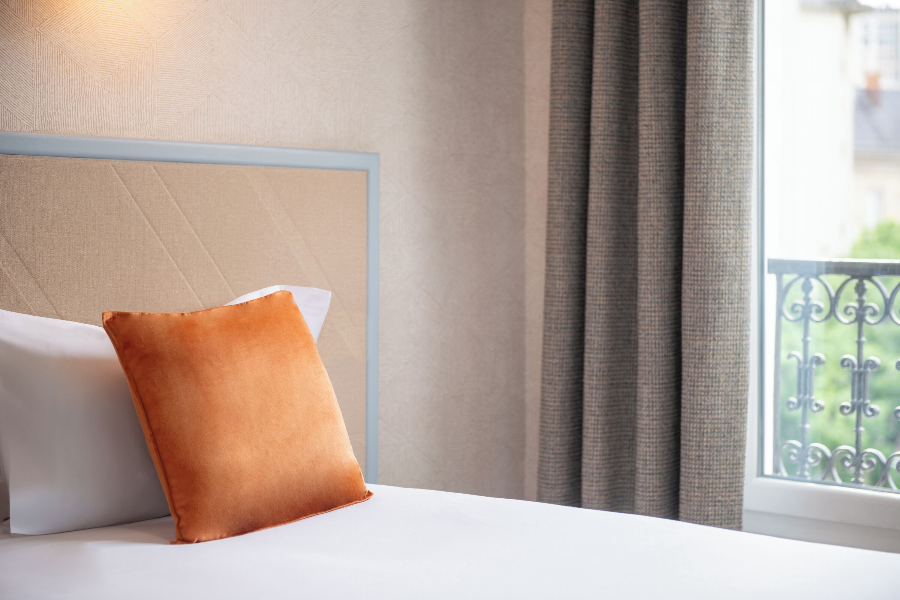 A neatly made bed with a white pillow and a single orange pillow, a light blue headboard, and a view of a balcony with a wrought iron railing.