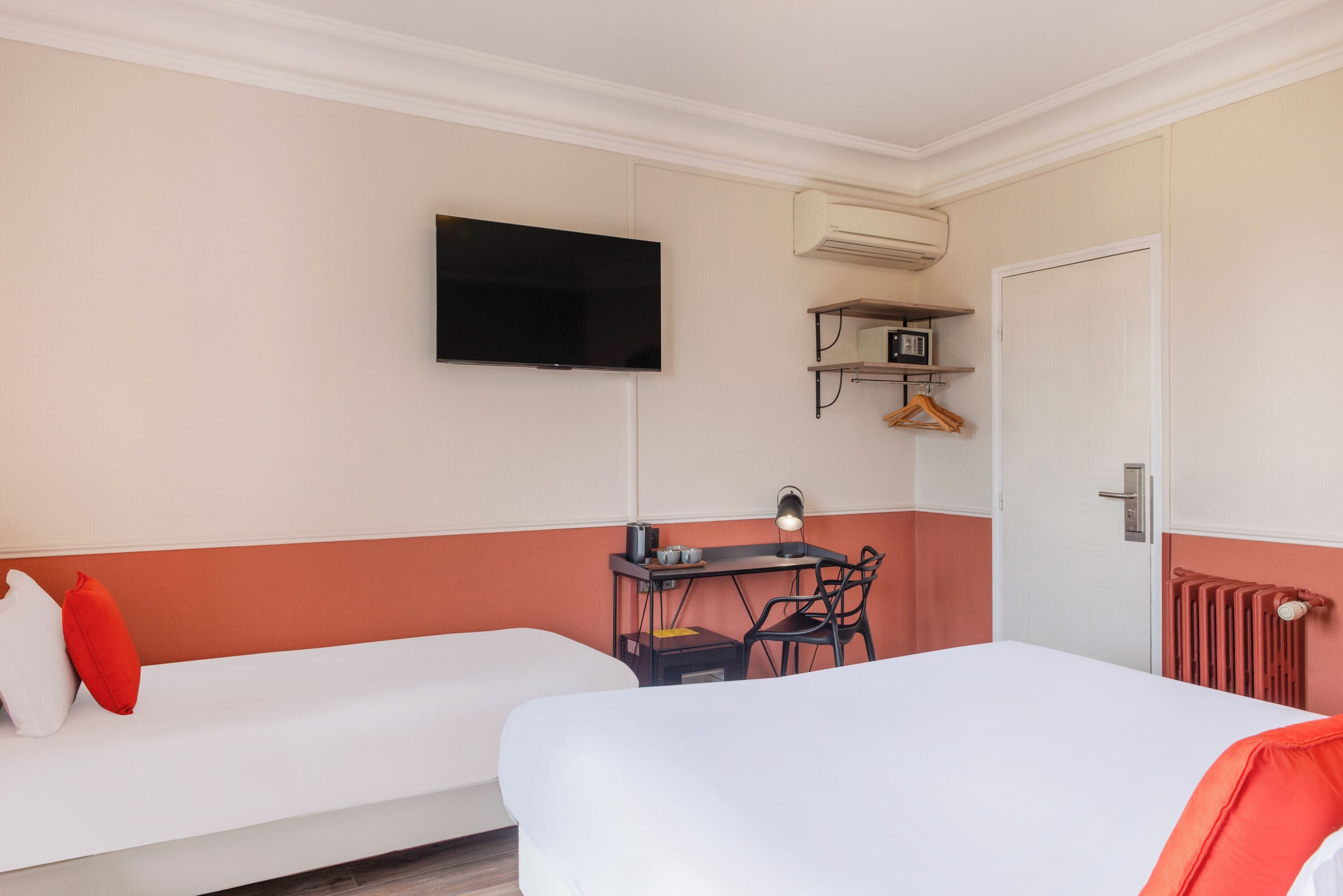 A hotel room with two beds, a TV, a desk with a chair, a radiator, and a coat rack.