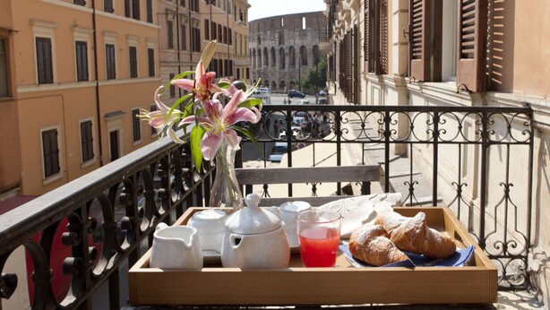 A balcony with a tray of breakfast items, including croissants and a vase of flowers, overlooking a historic building and a busy street. - Santi Quattro al Colosseo (Rome)