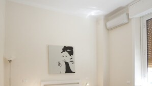 A bedroom with a bed, bedside tables, a floor lamp, a wall-mounted air conditioner, and a window with blinds. - Santi Quattro al Colosseo (Rome)