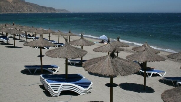 Una playa con filas de tumbonas y sombrillas de paja, un mar azul cristalino y un acantilado rocoso al fondo.