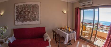 A room with a red sofa, a dining table with chairs, a large window with a view of the sea, and an air conditioning unit.