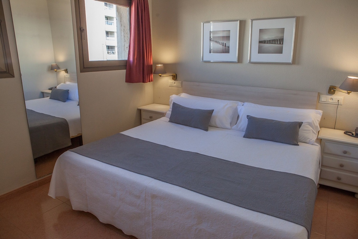 Una habitación de hotel con una cama grande, dos mesitas de noche con lámparas, un espejo y dos cuadros en la pared.