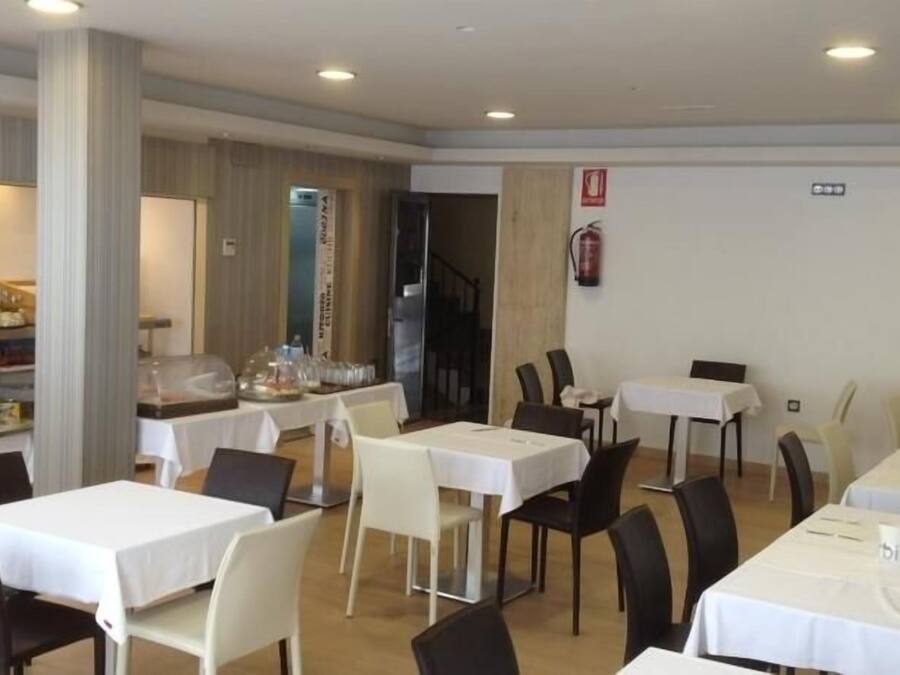 Un comedor con varias mesas y sillas, un área de cocina con refrigerador y encimera, y una escalera que sube a otro nivel.