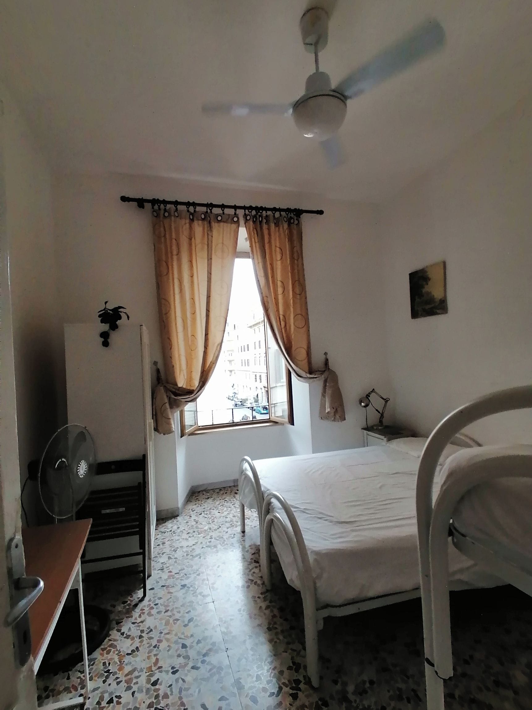 Un dormitorio con cama, ventilador de techo, ventana con cortinas, radiador y lámpara de pared.