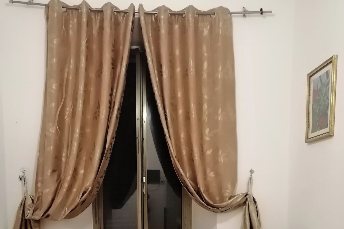 A room with a bed, a window with curtains, a wall-mounted picture, and a bedside table.