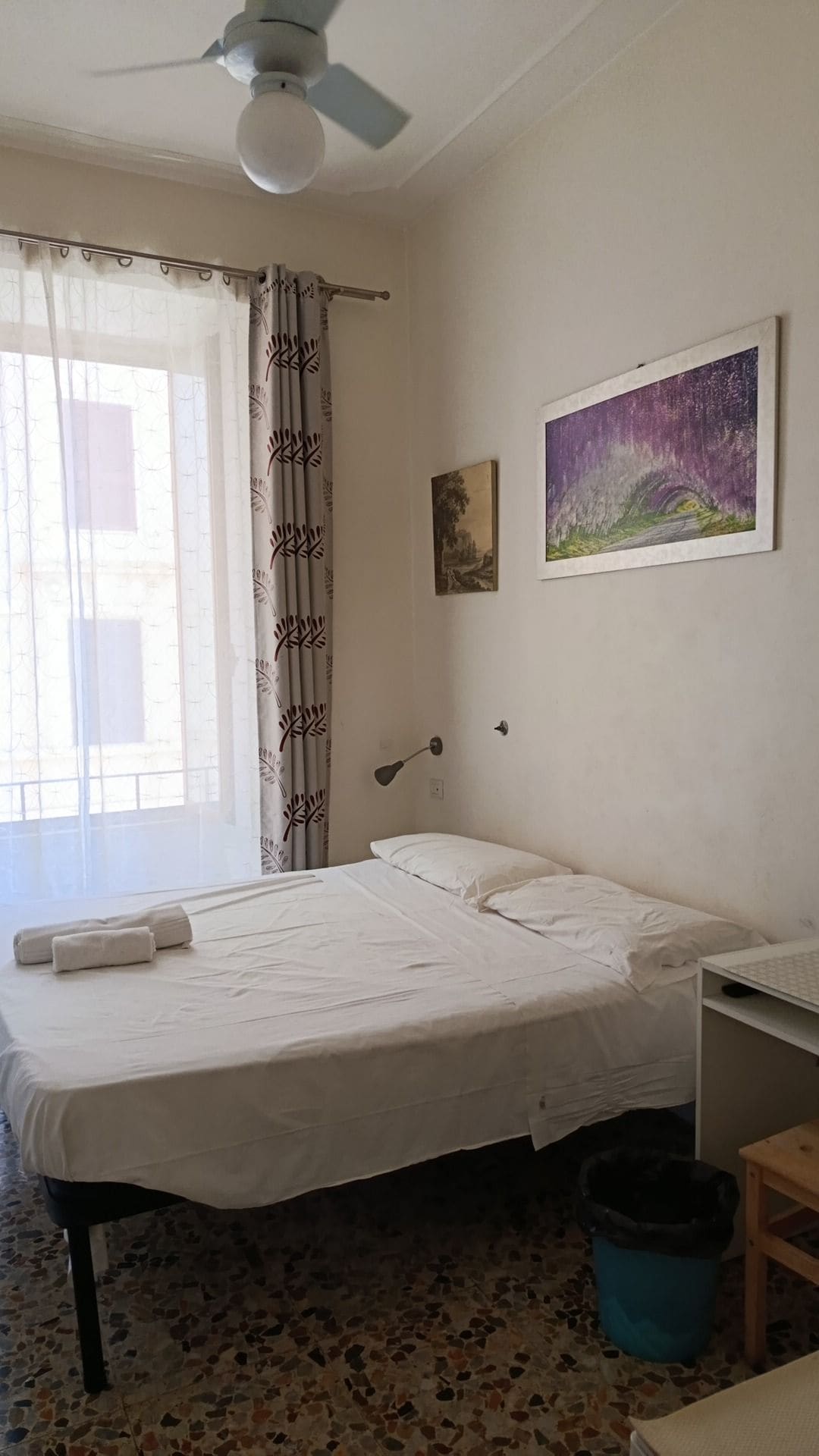 Un dormitorio con cama, escritorio, ventilador de techo, ventana con cortinas y un cuadro en la pared.