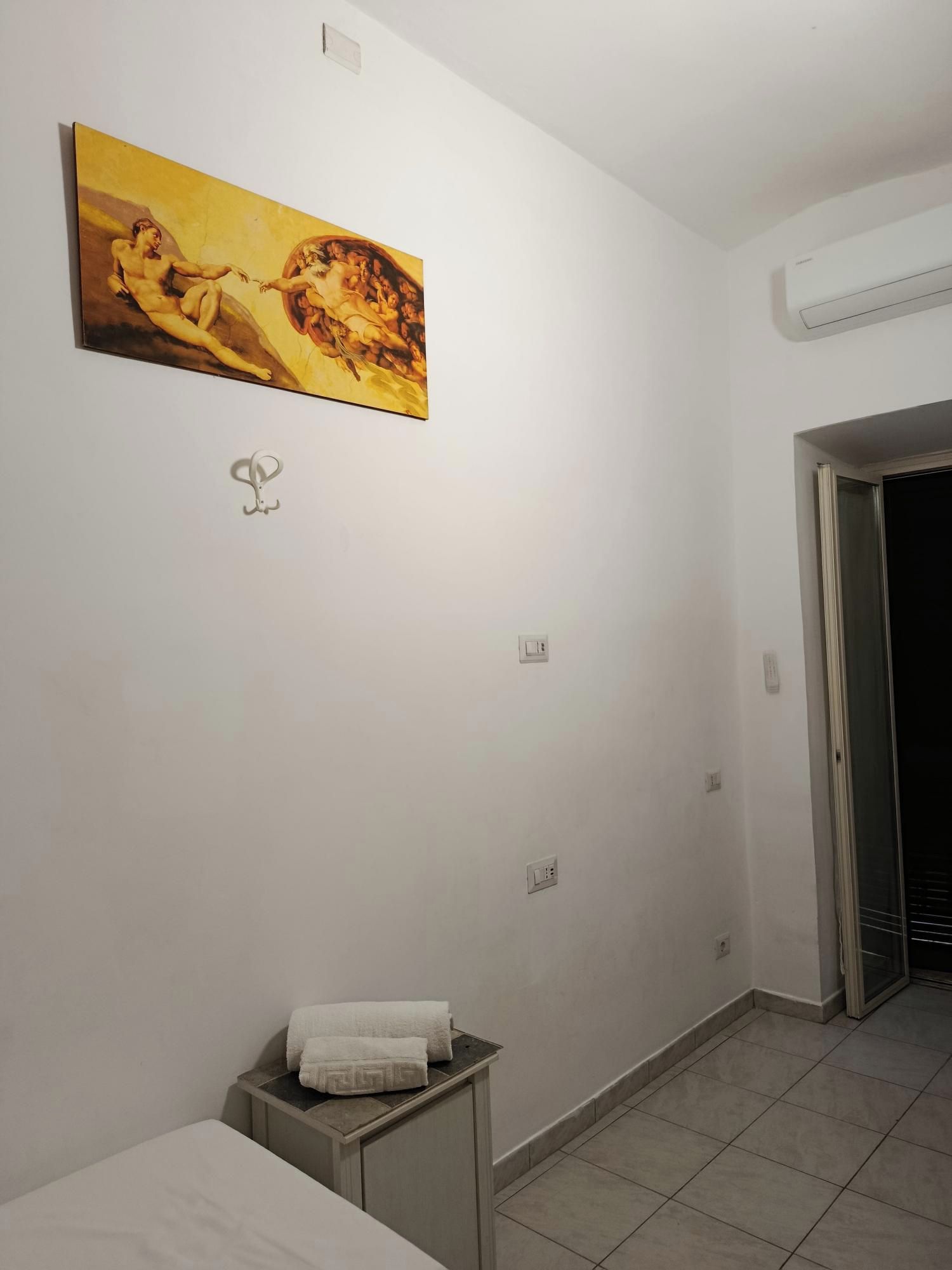 A hotel room with a bed, a small bedside table with a towel, an air conditioning unit, and a painting on the wall.