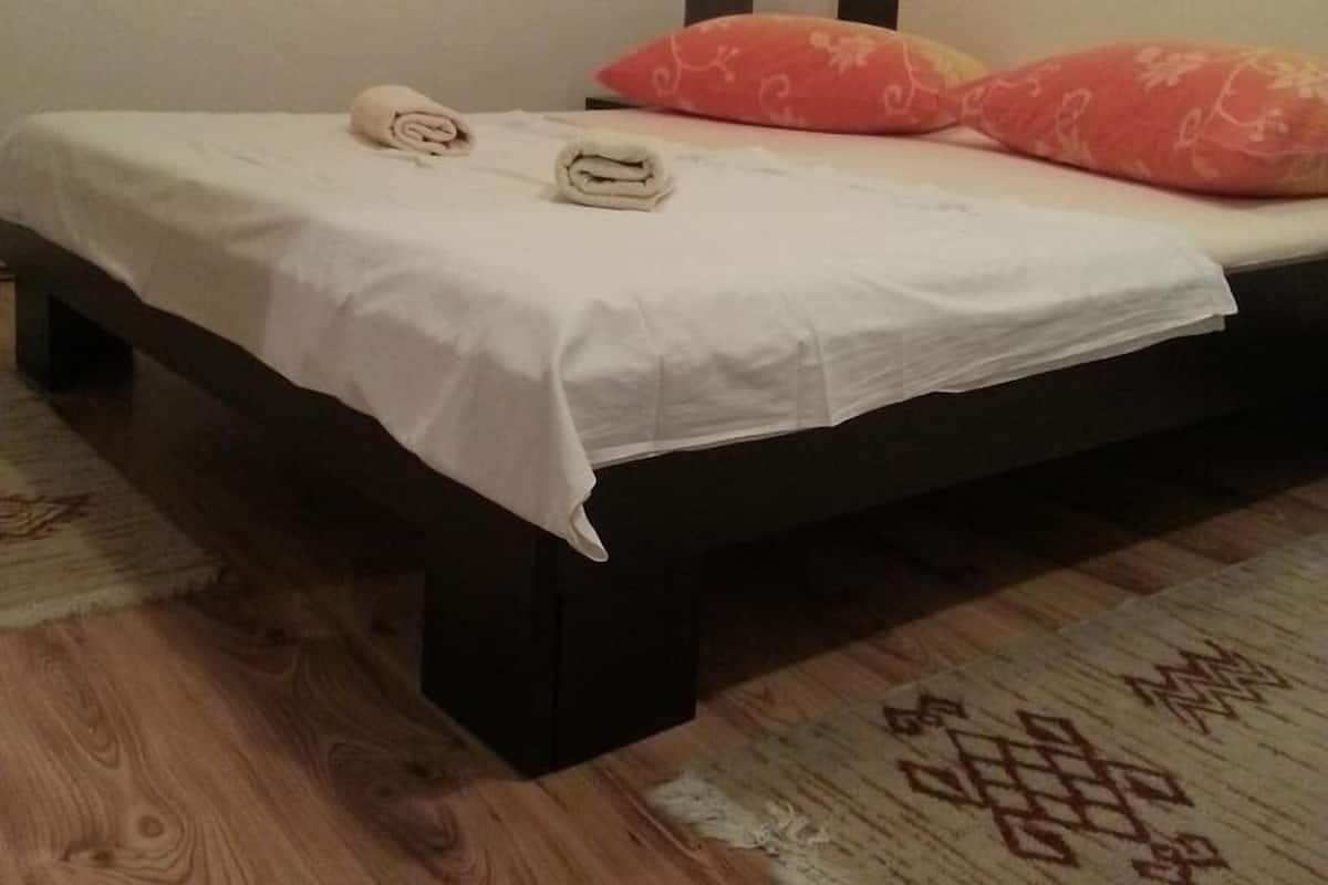 A single bed with a white bedspread, two rolled towels, and a red pillow in a room with a wooden floor and a patterned rug.