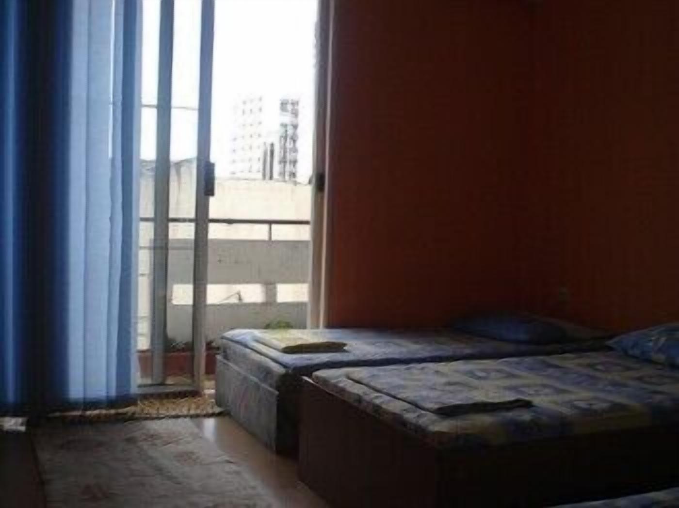 A small room with a bed, a window with curtains, and a view of buildings outside.