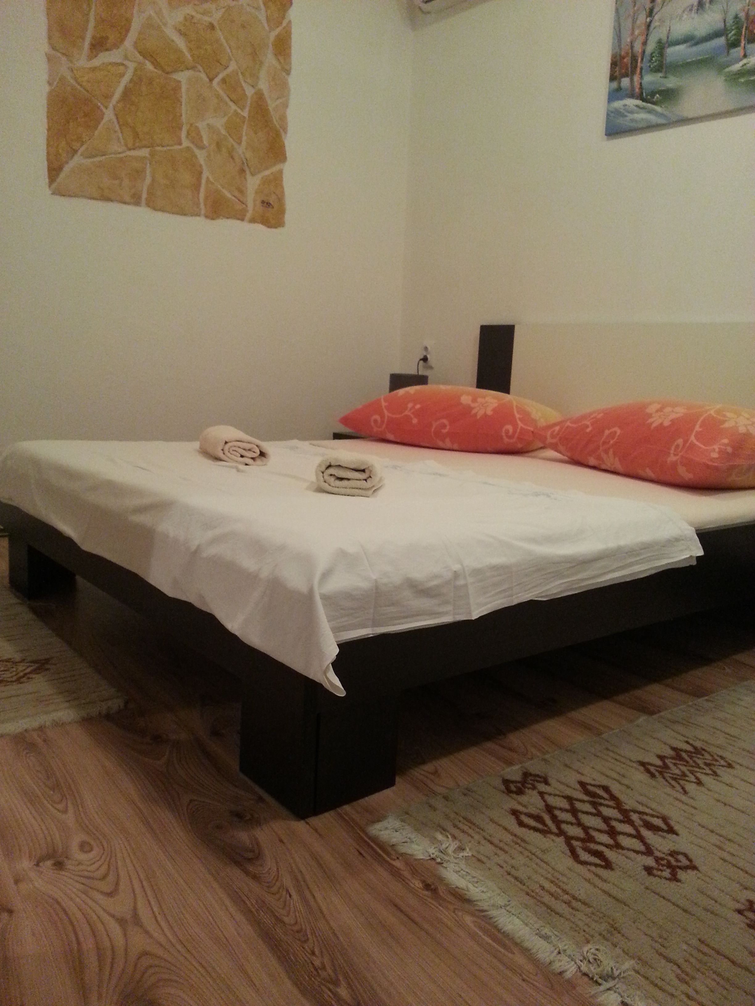 A single bed with a white sheet, two pillows, and a rolled towel. A stone wall and a painting are visible in the background.