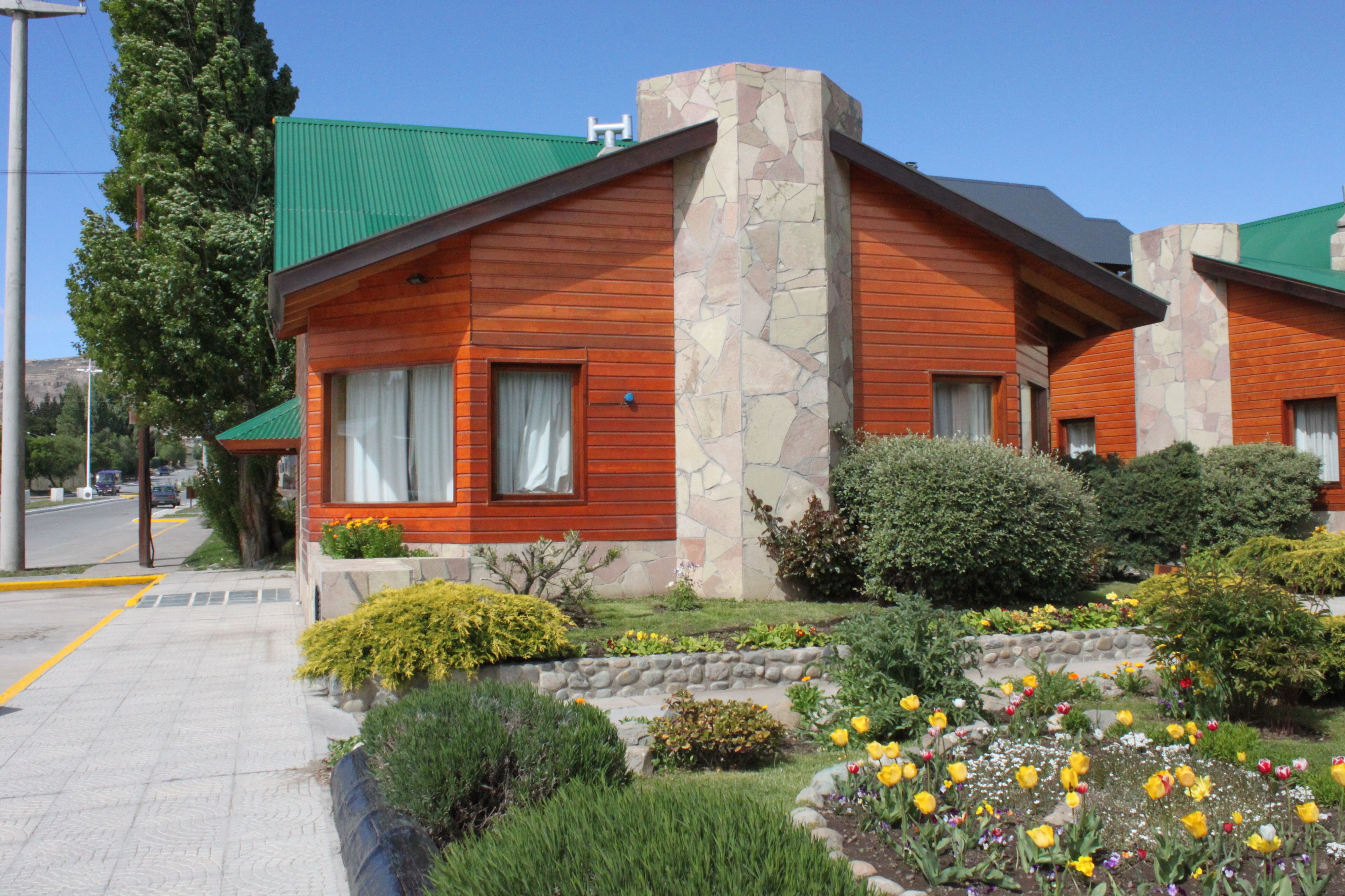 Une maison en bois à deux étages, dotée d’une cheminée en pierre, entourée d’un jardin bien entretenu où poussent différentes variétés de fleurs et d’arbustes.