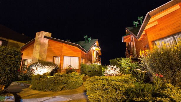 Une maison en bois avec une cheminée en pierre, entourée de jardins bien entretenus et bordée d’un sentier.
