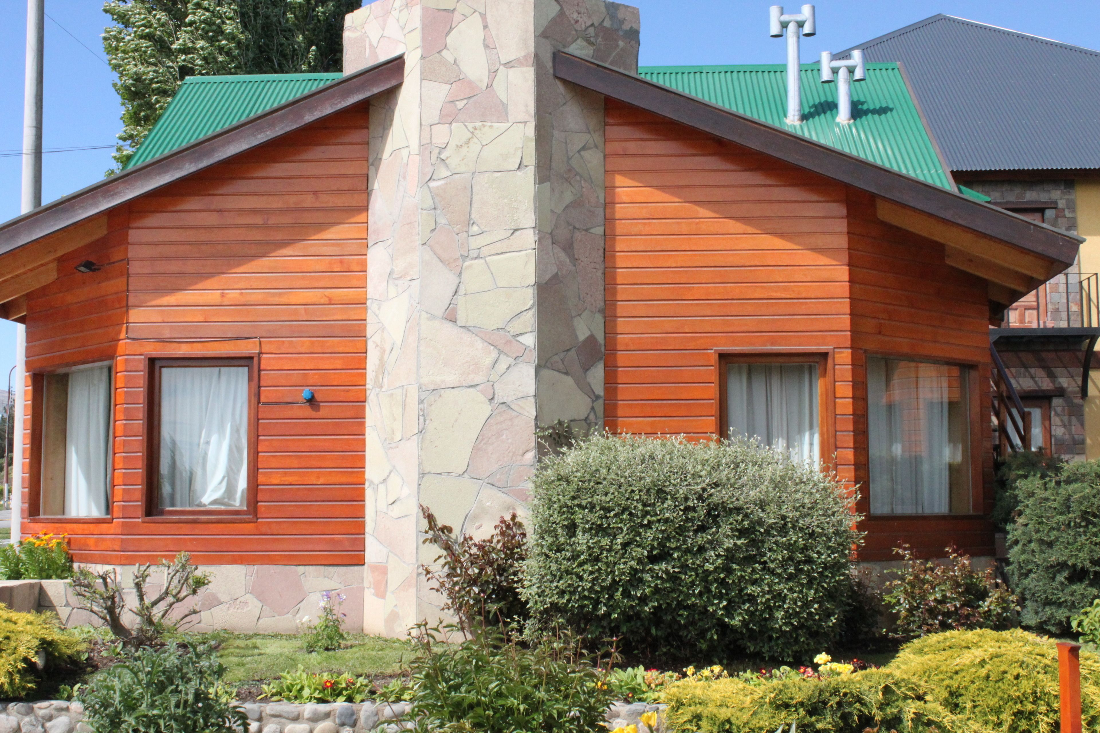 A two-story wooden house with a stone chimney, a green roof, and a well-maintained garden with various plants and flowers.