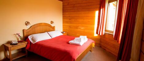 A neatly made bed with a red bedspread and white pillows, a wooden headboard, a nightstand with a lamp, and a window with curtains.