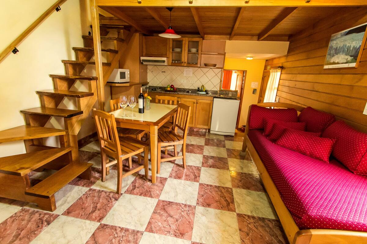 Interior acollidor d'una cabana amb zona de menjador, escala de fusta i un sofà vermell.