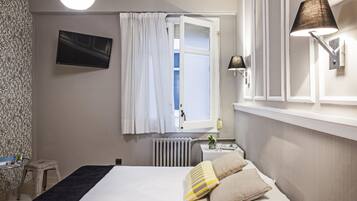 Een slaapkamer met een bed, nachtkastje, lamp, raam met gordijnen, radiator en een televisie aan de muur.