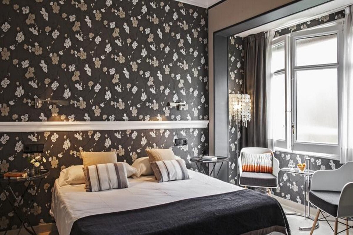 A hotel room with a large bed, a small table, a chair, and a window with curtains.