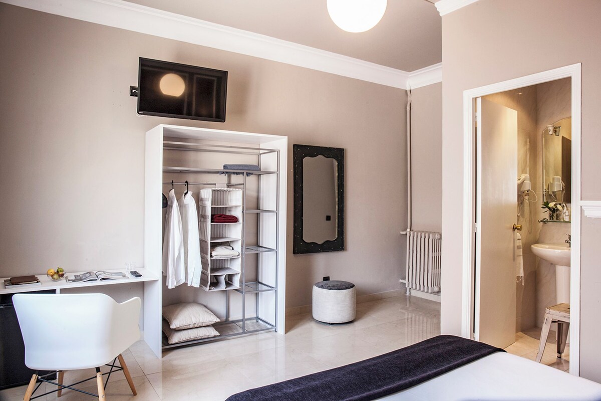 A modern bedroom with a walk-in closet, a bedside table, a chair, a round ottoman, and a bathroom with a sink and mirror.