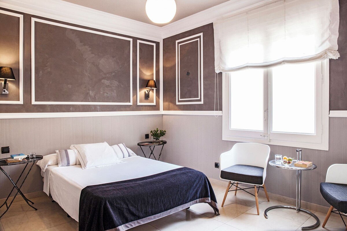 A hotel room with a bed, a desk, a chair, a small table, and a window with curtains.
