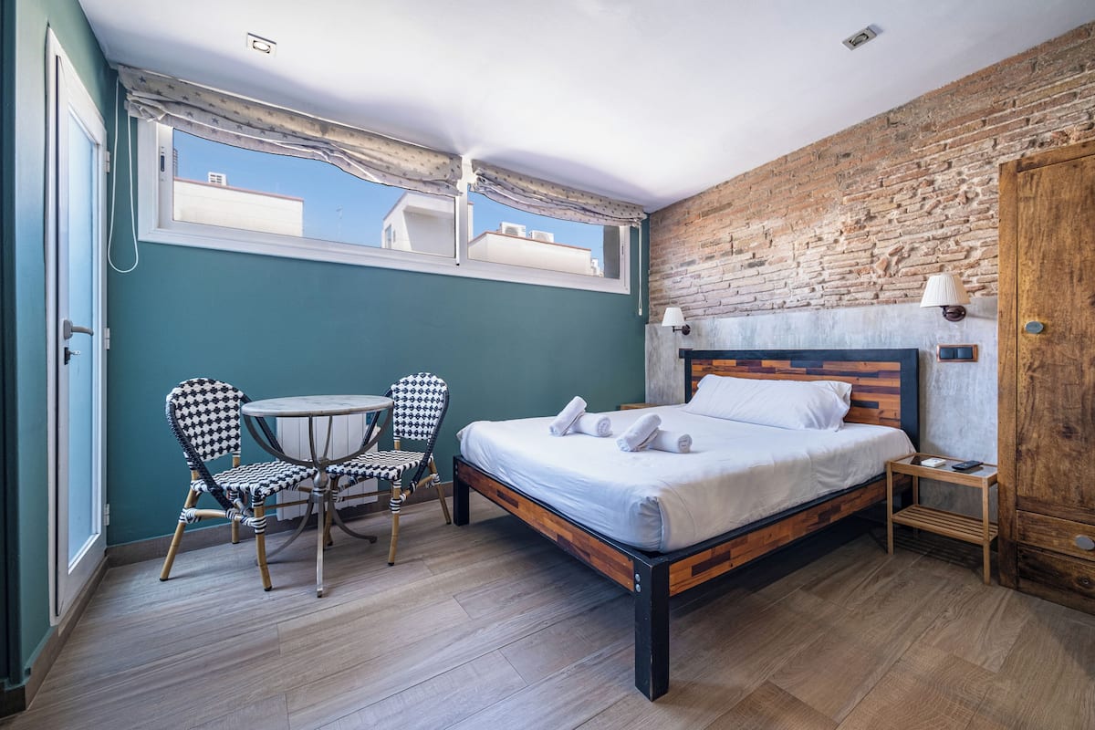 Ein Schlafzimmer mit Bett, Sessel, Nachttisch und Fenster mit Blick auf Gebäude.