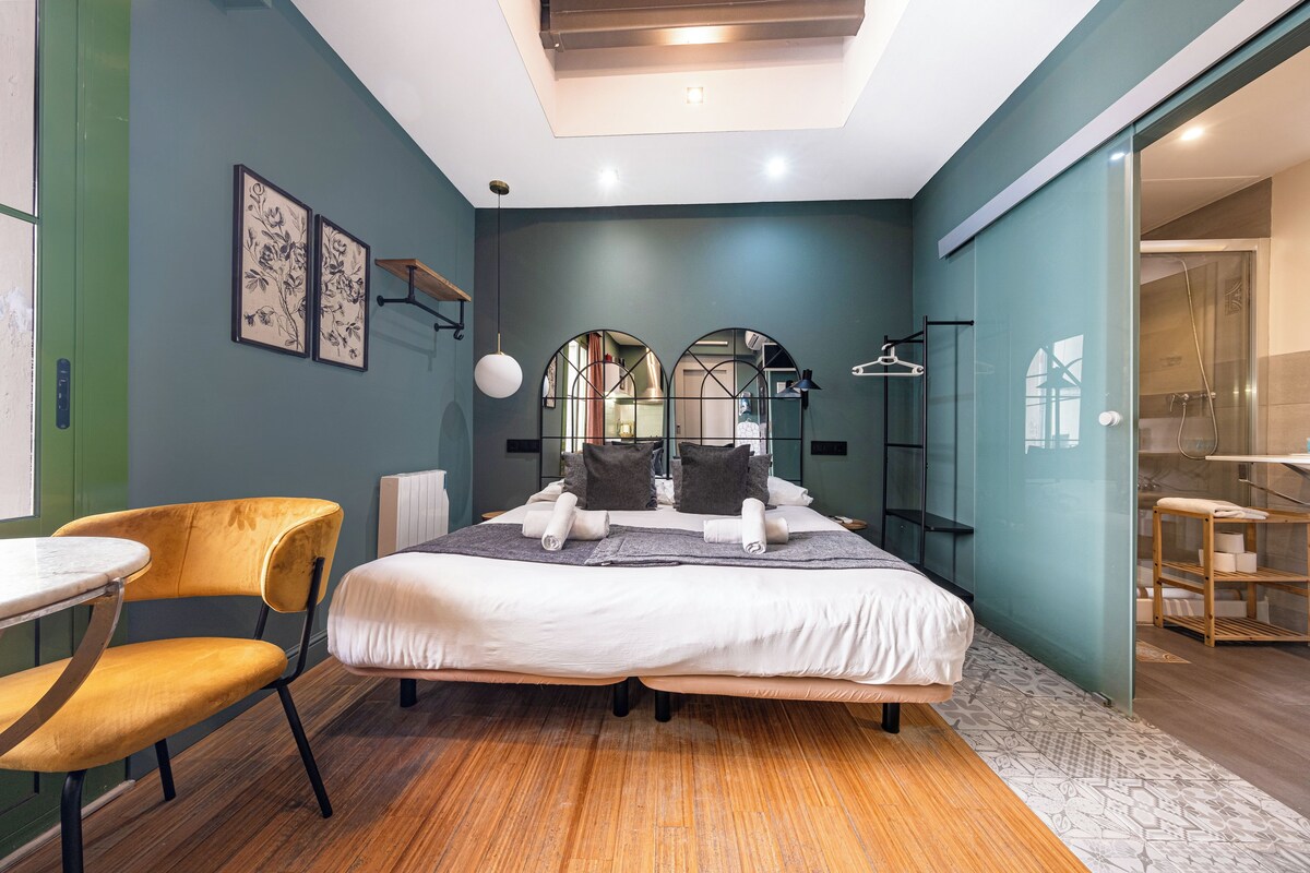 Una habitación de hotel moderna con una cama grande, una mesita con una silla y un baño visible a través de una puerta de cristal.