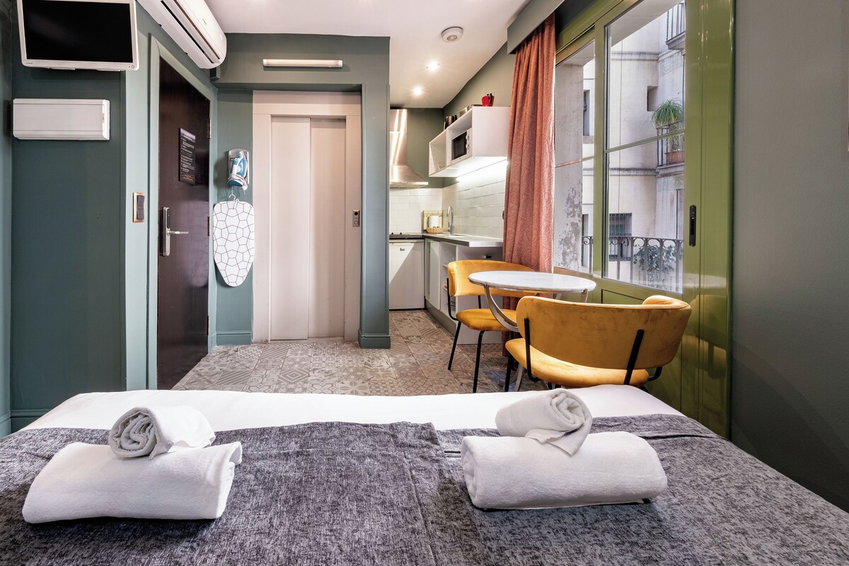 Habitación de hotel con cama, kitchenette, mesa de comedor con sillas y vista al balcón exterior.