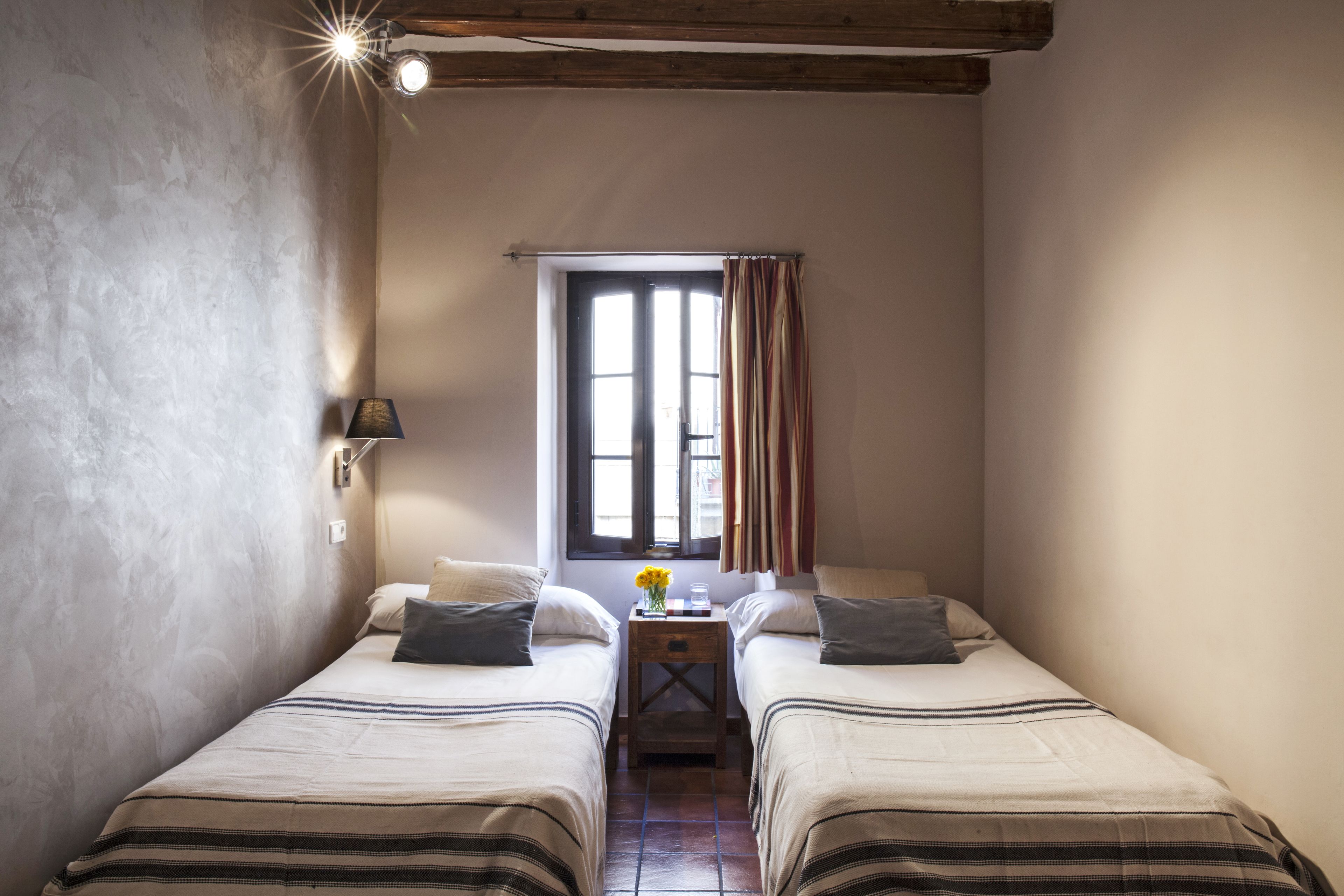 Una habitación pequeña y acogedora con dos camas, una ventana con cortinas, una mesita de noche con lámpara y una lámpara de pared.
