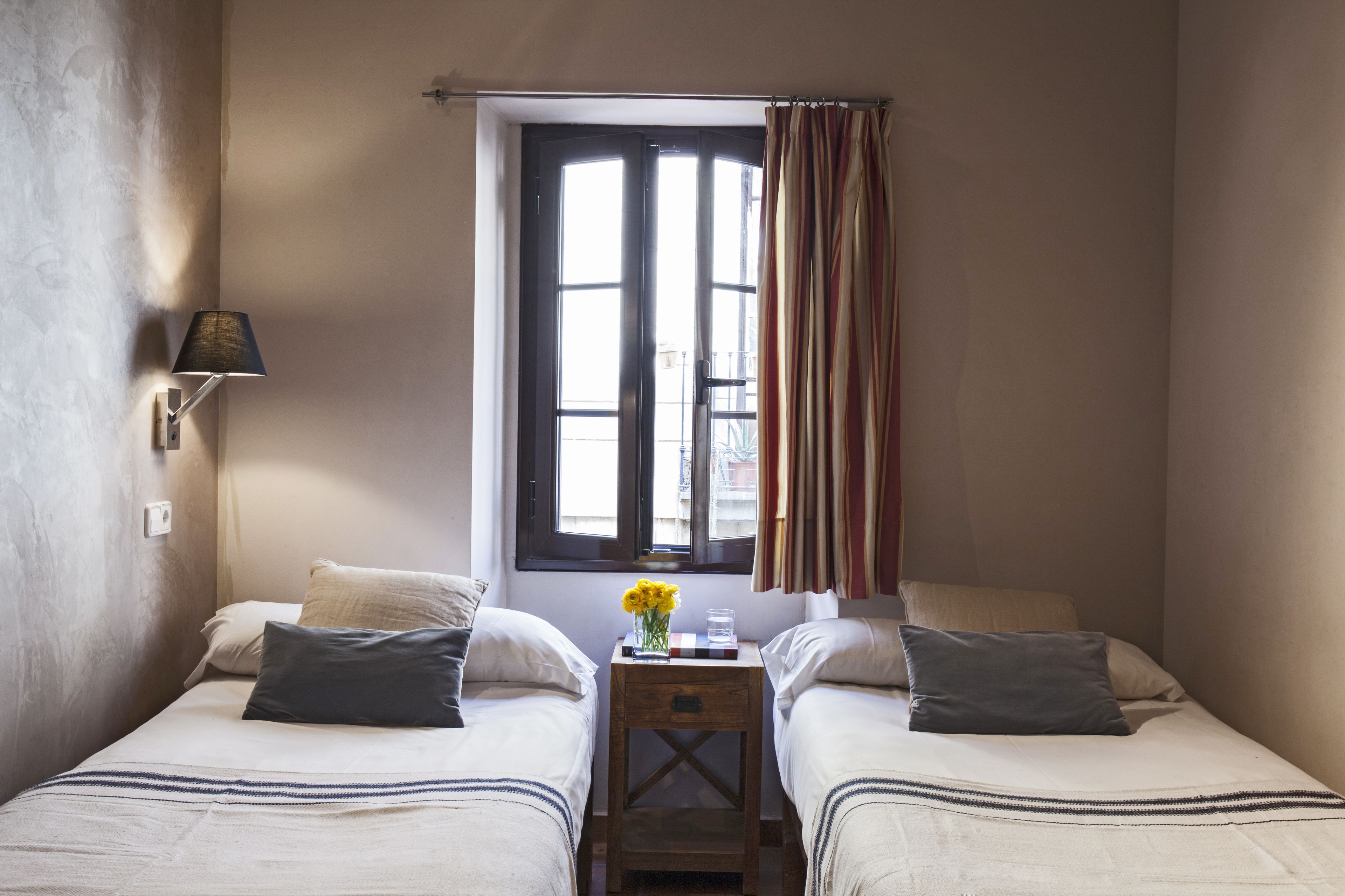 Une petite chambre bien éclairée, avec deux lits, une fenêtre avec des rideaux, une lampe de chevet et une petite table sur laquelle est posé un vase de fleurs.