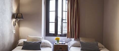 A small, well-lit bedroom with two beds, a window with curtains, a bedside lamp, and a small table with a vase of flowers.